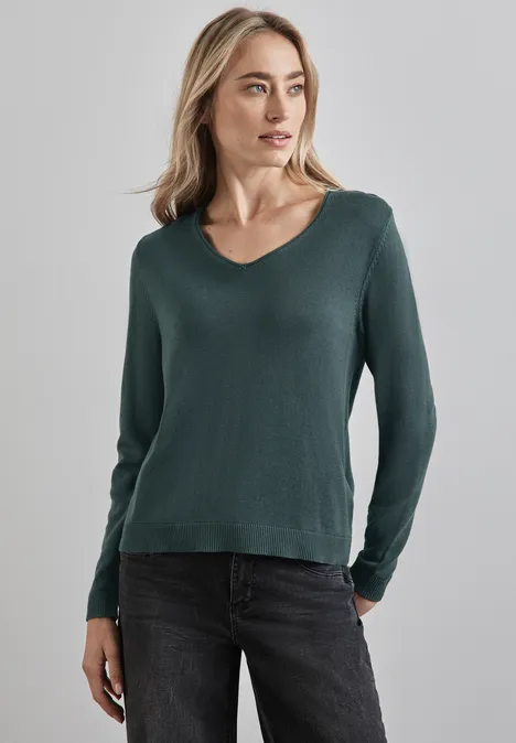 Frau mit grünem Pullover und dunkler Jeans steht vor grauem Hintergrund.